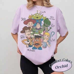 May include: Orchid-colored t-shirt featuring a graphic of Toy Story characters, including Woody, Buzz Lightyear, and Mr. Potato Head. The shirt has a Comfort Colors label.