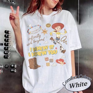 May include: A white t-shirt featuring a graphic print of a cowgirl character, a brown cowboy boot, alphabet blocks, a sheriff star, and a beaded necklace. The text reads infinity and beyond, I KNEW IT, and I KNEW YOU in various colors.