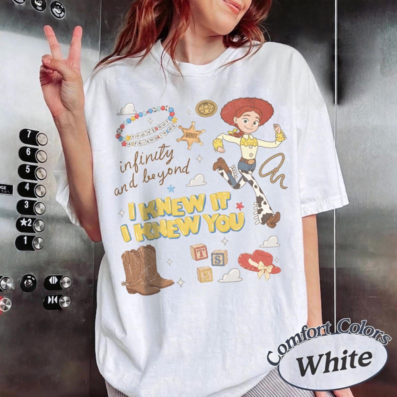 May include: A white t-shirt featuring a graphic print of a cowgirl character, a brown cowboy boot, alphabet blocks, a sheriff star, and a beaded necklace. The text reads infinity and beyond, I KNEW IT, and I KNEW YOU in various colors.