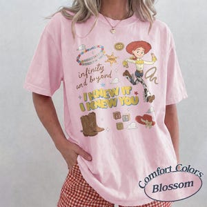 May include: A light pink t-shirt featuring a graphic print of the character Jessie from Toy Story, brown cowboy boots, a red hat, alphabet blocks, and a beaded necklace. The text reads infinity and beyond, I KNEW IT, and I KNEW YOU.