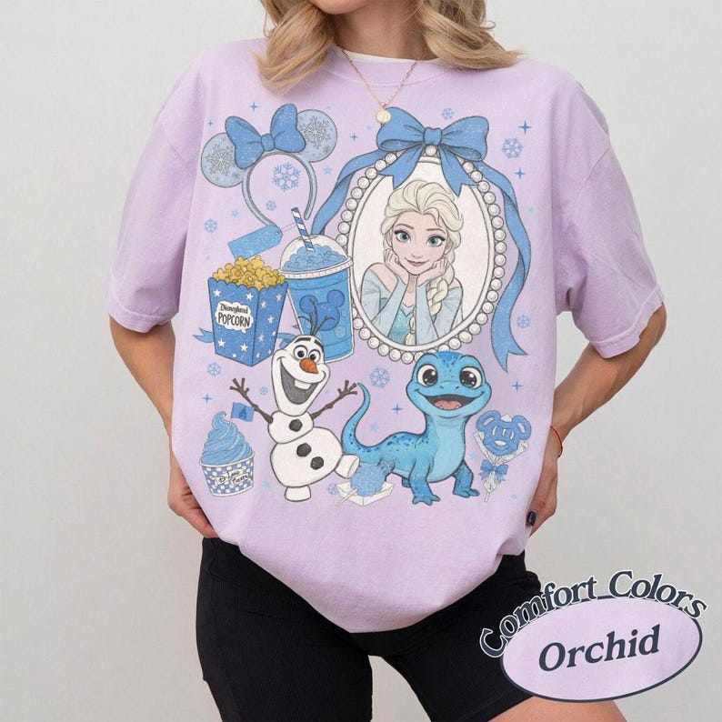 Op de afbeelding: Orchid Comfort Colors T-shirt met een afbeelding van Elsa uit Frozen, Olaf en Bruni de salamander. Het ontwerp bevat een blauwe strik, popcorn, een drankje en een cupcake. Het shirt heeft korte mouwen.