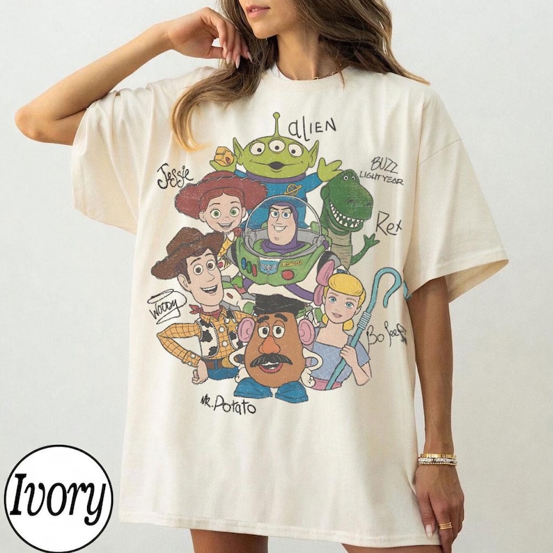 May include: Ivory-colored t-shirt featuring a colorful graphic of characters from the animated film Toy Story. The design includes Woody, Buzz Lightyear, Jessie, Rex, Mr. Potato Head, Bo Peep, and an alien. The word "Ivory" is printed in a circle at the bottom.
