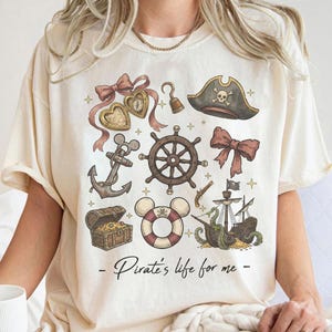 Könnte beinhalten: Cremefarbenes T-Shirt mit einem Piraten-Motiv. Das Design umfasst einen Piratenhut, ein Steuerrad, einen Anker, eine Schatzkiste und eine Schwimmweste mit Totenkopf. Der Text "- Pirate's life for me -" ist darunter gedruckt.