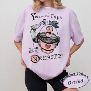 Puede incluir: Camiseta Comfort Colors color orqu&iacute;dea con una ilustraci&oacute;n de dibujos animados de un personaje en un cubo rosa y el texto "You See the Hat?! I Am Mrs. NESBITT!" en negro. El dise&ntilde;o incluye un sombrero azul y un coraz&oacute;n.