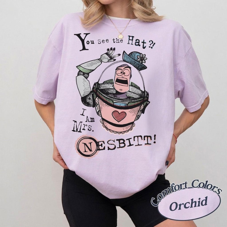 Puede incluir: Camiseta Comfort Colors color orqu&iacute;dea con una ilustraci&oacute;n de dibujos animados de un personaje en un cubo rosa y el texto "You See the Hat?! I Am Mrs. NESBITT!" en negro. El dise&ntilde;o incluye un sombrero azul y un coraz&oacute;n.