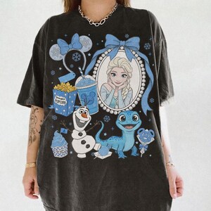 Op de afbeelding: Zwart T-shirt met een afbeelding van Elsa uit Frozen, omringd door blauwe strikken, sneeuwvlokken en thema-elementen. Bevat afbeeldingen van Olaf, een blauwe hagedis, popcorn en een drankje. Het shirt heeft een vintage uitstraling.