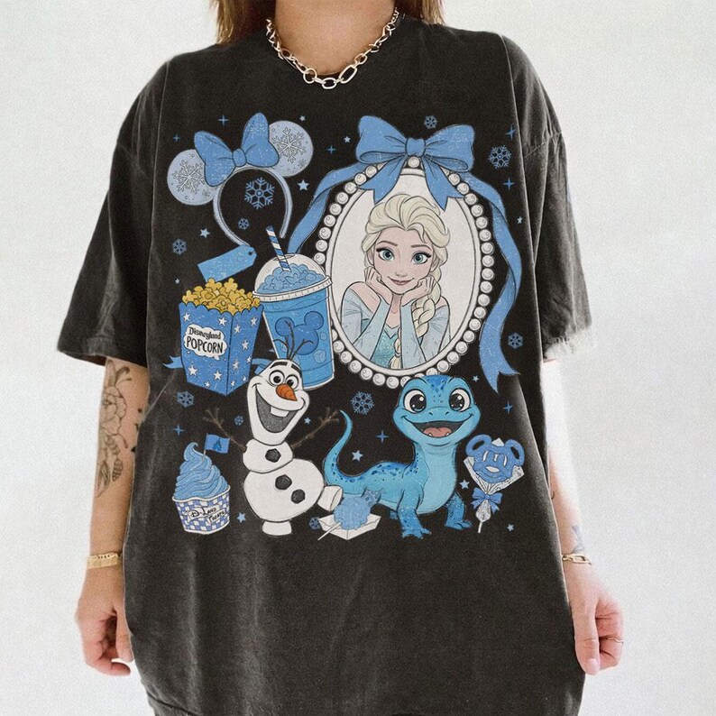 Op de afbeelding: Zwart T-shirt met een afbeelding van Elsa uit Frozen, omringd door blauwe strikken, sneeuwvlokken en thema-elementen. Bevat afbeeldingen van Olaf, een blauwe hagedis, popcorn en een drankje. Het shirt heeft een vintage uitstraling.
