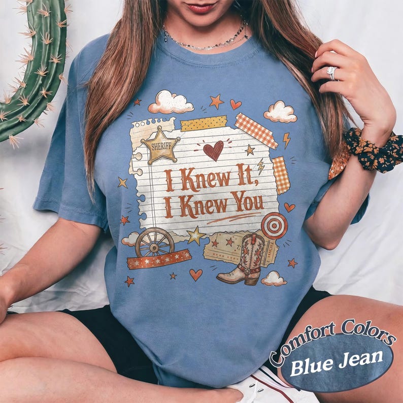 May include: A blue jean coloured t-shirt featuring a graphic of a lined notepad with the text I Knew It, I Knew You. The design includes a sheriff star, a heart, a wagon wheel, a cowboy boot, and various small stars and clouds in warm tones.