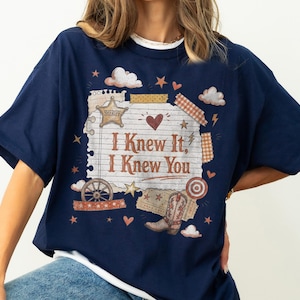 May include: A navy blue t-shirt featuring a graphic print of a lined paper sheet with the text "I Knew It, I Knew You". The design includes a sheriff star, a cowboy boot, a wagon wheel, clouds, hearts, stars, and decorative tape strips.
