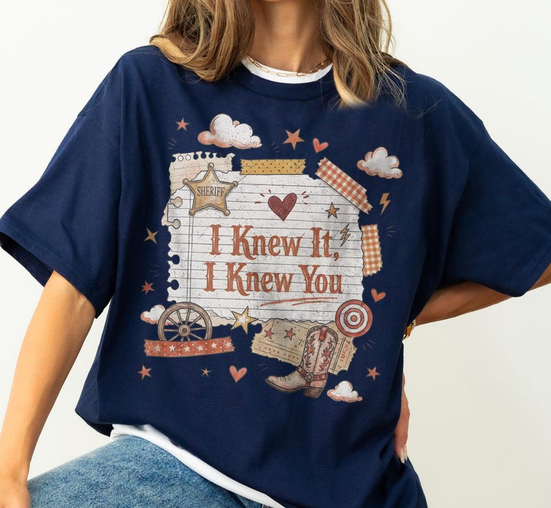 May include: A navy blue t-shirt featuring a graphic print of a lined paper sheet with the text "I Knew It, I Knew You". The design includes a sheriff star, a cowboy boot, a wagon wheel, clouds, hearts, stars, and decorative tape strips.
