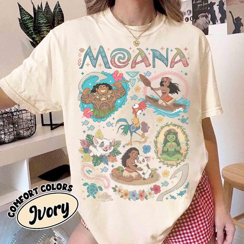 May include: Cream-coloured t-shirt with the word "MOANA" in colourful letters. The shirt features cartoon illustrations of characters and scenes from the film, including Maui, Moana, and Pua. The shirt also has the words "COMFORT COLORS" and "Ivory" printed on it.