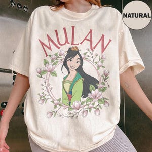 May include: A cream-colored short-sleeve t-shirt featuring a graphic of the character Mulan in a green and yellow outfit. The design includes the text MULAN in red letters above the character, surrounded by a circular frame of pink magnolia flowers.