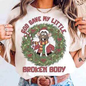 May include: Cream-colored t-shirt featuring a cartoon rat in a Santa hat and scarf, holding a candy cane within a Christmas wreath. The text "GOD SAVE MY LITTLE BROKEN BODY" is arched above and below the wreath.