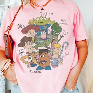 May include: Pink t-shirt featuring a graphic of Toy Story characters, including Woody, Buzz Lightyear, and Mr. Potato Head. The names of the characters are written above them in black. The shirt has short sleeves and a crew neck.