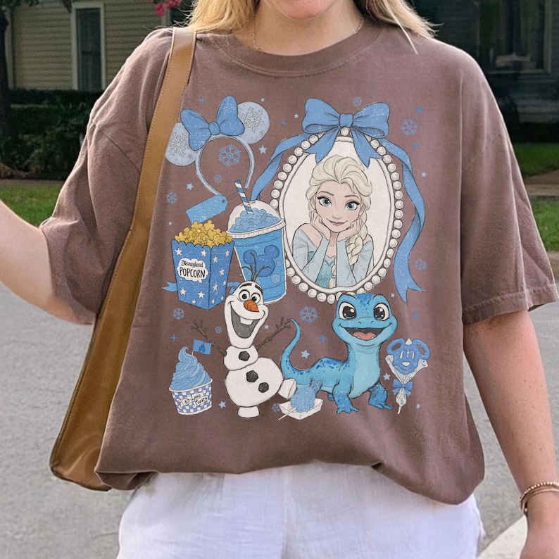 Op de afbeelding: Bruin T-shirt met een afbeelding van Elsa, Olaf en Bruni uit Frozen. Het ontwerp bevat een blauwe strik, sneeuwvlokken en een popcornbox. Het shirt heeft een relaxte pasvorm.