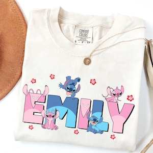 May include: Cream-colored t-shirt featuring the name "EMILY" in pink and blue ombre letters, with cartoon characters and red flower accents. A gold necklace with a pendant is visible.
