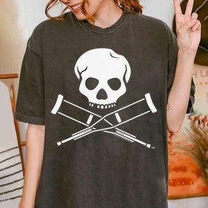 May include: A black t-shirt featuring a white skull and crossbones design. The skull has a white outline and the crossbones are made of crutches.