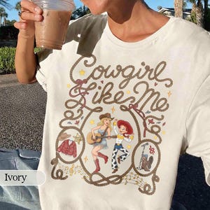 May include: An ivory t-shirt featuring a graphic print with the text Cowgirl like Me in rope-style lettering. The design includes illustrations of two figures, a guitar, a red patterned jacket, a cowboy hat, and boots, all framed by a rope border.