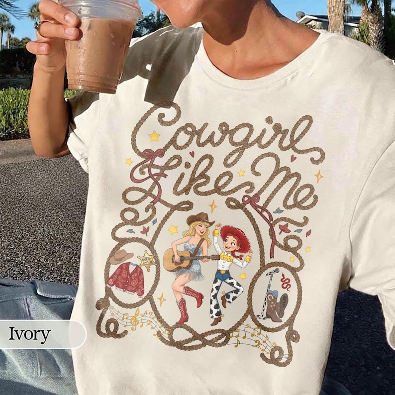 May include: An ivory t-shirt featuring a graphic print with the text Cowgirl like Me in rope-style lettering. The design includes illustrations of two figures, a guitar, a red patterned jacket, a cowboy hat, and boots, all framed by a rope border.