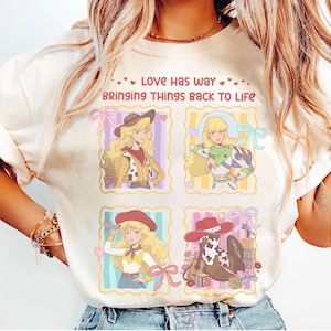 May include: A cream-colored t-shirt featuring a graphic print with four square panels. The text reads, "LOVE HAS WAY BRINGING THINGS BACK TO LIFE." The panels depict a blonde character in various outfits, including a cowgirl and space ranger, plus a cowboy boot.