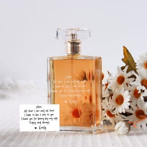 May include: A clear glass bottle of perfume with a white label that reads "Mom All that I am and all that I hope to be, I owe to you. Thank you for being by my side Today and always Emily". The bottle is sitting on a white surface with a bouquet of white daisies in the background.