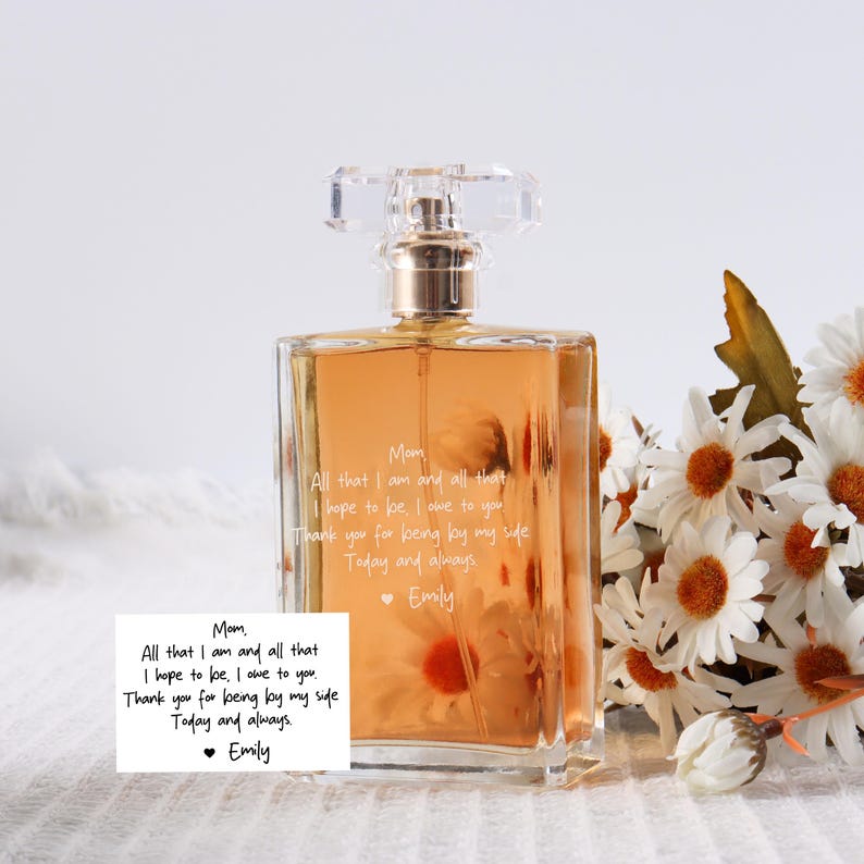 May include: A clear glass bottle of perfume with a white label that reads "Mom All that I am and all that I hope to be, I owe to you. Thank you for being by my side Today and always Emily". The bottle is sitting on a white surface with a bouquet of white daisies in the background.