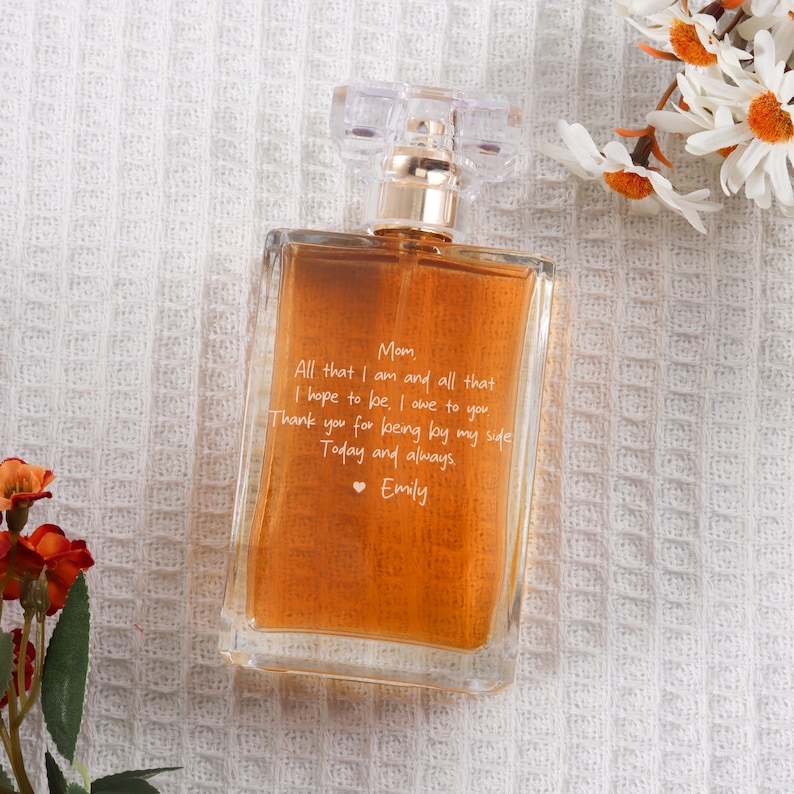 May include: A clear glass bottle of perfume with a gold cap. The bottle is filled with a brown liquid and has a white inscription that reads: "Mom, All that I am and all that I hope to be, I owe to you. Thank you for being by my side. Today and always. &bull; Emily"