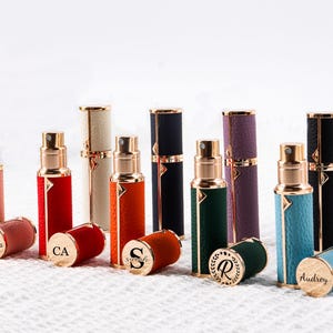 May include: A collection of personalized perfume atomizers in various colors, including pink, red, orange, green, purple, black, and teal. Each atomizer has a gold-colored top and a matching colored base with a name or initial engraved.
