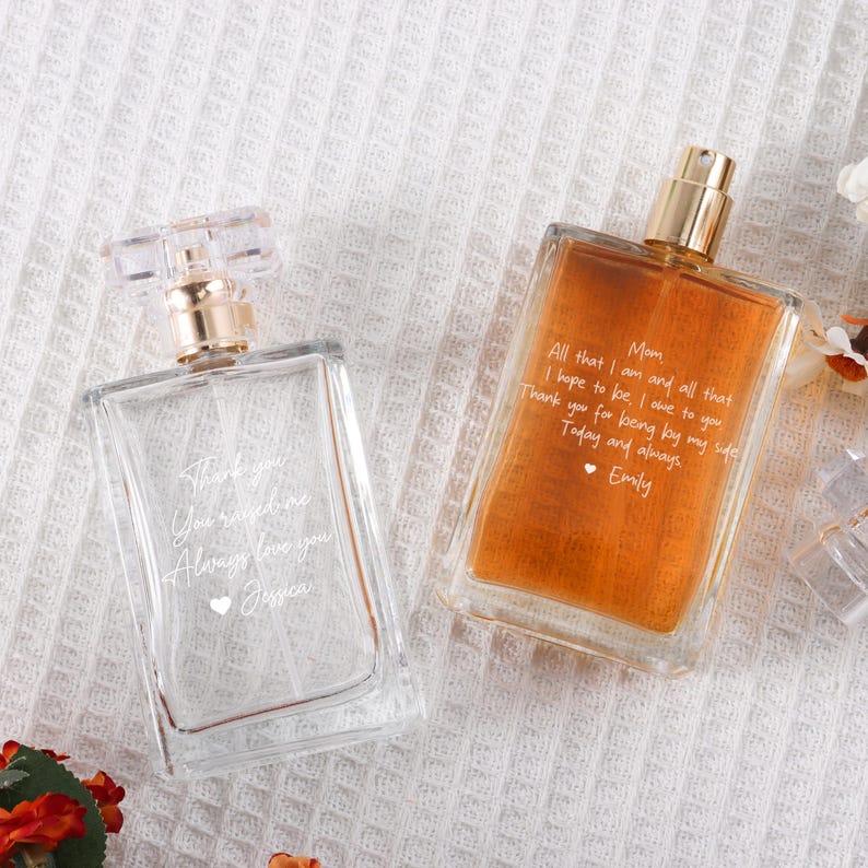 May include: Two clear glass perfume bottles with gold caps. The bottle on the left has the text "Thank you you mean the world Always love you Jessica" etched on it. The bottle on the right has the text "Mom All that I am and all that I hope to be, I owe to you Thank you for being by my side Today and always, Emily" etched on it.
