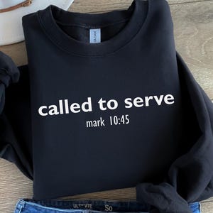 Könnte beinhalten: Schwarzes Sweatshirt mit dem weißen Text "called to serve" und "mark 10:45". Das Sweatshirt ist gefaltet, mit einer Jeans und einem weißen Hut im Hintergrund. Das Sweatshirt besteht aus einem weichen Material.