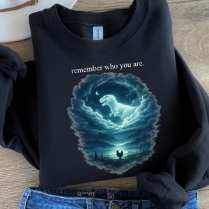 Puede incluir: Sudadera negra con el texto "remember who you are". El gráfico presenta un dinosaurio brillante dentro de un óvalo lleno de nubes con una luna creciente. Una gallina está en primer plano. La sudadera está doblada sobre unos vaqueros azules.