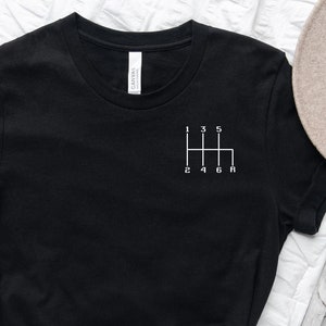 Könnte beinhalten: Schwarzes T-Shirt mit einem weißen Aufdruck eines Schaltmusters für ein Auto. Die Zahlen 1, 2, 3, 4, 5 und R sind auf dem Aufdruck sichtbar.