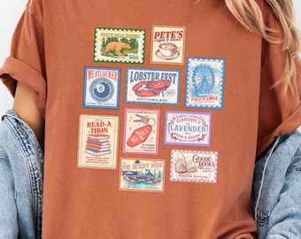 May include: A rust-colored t-shirt featuring a collection of vintage-style postage stamp designs. The stamps showcase various themes, including a bear, coffee, a lobster, and books. The shirt is paired with a denim jacket.