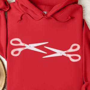 Könnte beinhalten: Ein leuchtend roter Kapuzenpullover mit einer weißen Scheren-Grafik. Die Schere ist im Cut-out-Stil dargestellt, wobei die Klingen getrennt sind. Der Hoodie hat eine Kapuze mit Kordelzug und eine Vordertasche.