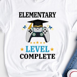 May include: White t-shirt featuring the text "ELEMENTARY LEVEL COMPLETE" in black and blue. A graduation cap sits above a grey and black video game controller. The design includes gold stars and blue diploma graphics.
