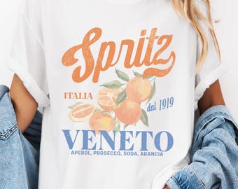 May include: White t-shirt with an orange "Spritz" graphic, featuring oranges and text that reads "Italia Veneto dal 1919". The shirt also lists ingredients: Aperol, Prosecco, Soda, Arancia.