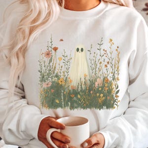 Puede incluir: Sudadera blanca con un diseño caprichoso de fantasma en medio de un campo de flores. El fantasma tiene una cara sencilla y amigable. Los elementos florales son de varios colores, creando una estética suave y natural. Una prenda de vestir acogedora y de temporada.