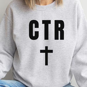 Op de afbeelding: Een lichtgrijs sweatshirt met de zwarte tekst "CTR" boven een zwart kruis. De sweatshirt heeft een ronde hals en lijkt gemaakt te zijn van een zacht materiaal. De persoon draagt een blauwe jeans.