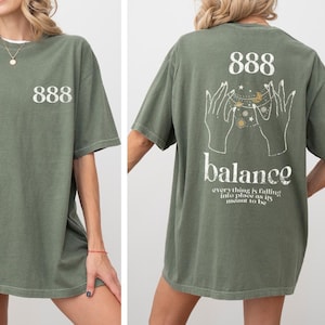 May include: Olive green t-shirt with the number 888 printed on the front and back. The back of the shirt features a graphic of hands, celestial elements, and the word "balance" with the phrase "everything is falling into place as it's meant to be."