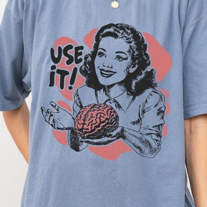 Puede incluir: Una camiseta azul con una ilustración retro de una mujer sosteniendo un cerebro. El texto "USE IT!" está impreso en la camiseta.