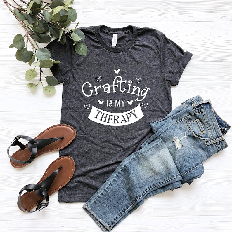 May include: A dark grey t-shirt with white text that reads "Crafting is my therapy" with small hearts around the text. The shirt is laid flat on a white wooden surface with a pair of brown sandals and blue jeans.