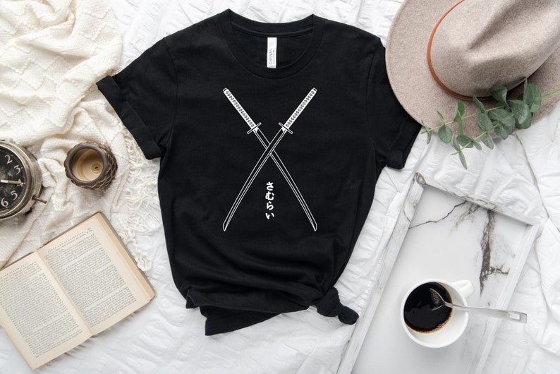 Może przedstawiać: Czarny T-shirt z białym nadrukiem dw&oacute;ch skrzyżowanych mieczy i japońskim tekstem "さむらい" (samuraj).