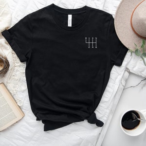 May include: Black t-shirt with a white graphic of a 6-speed manual transmission gear shift pattern.