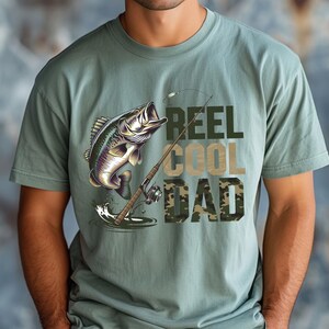 May include: Sage green t-shirt featuring a graphic of a leaping fish, fishing rod, and the words "REEL COOL DAD" in a mix of green and camouflage lettering. The shirt is a casual style.