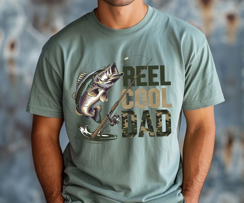 May include: Sage green t-shirt featuring a graphic of a leaping fish, fishing rod, and the words "REEL COOL DAD" in a mix of green and camouflage lettering. The shirt is a casual style.