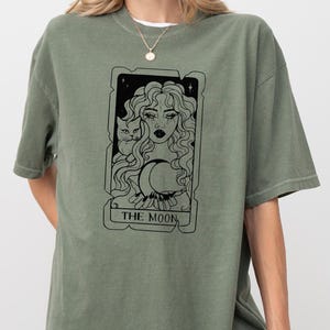 May include: A green t-shirt with a black and white illustration of a tarot card depicting "The Moon". The card features a woman with long hair, a cat, and a crescent moon.