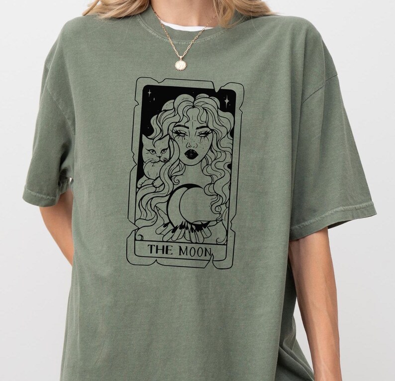 May include: A green t-shirt with a black and white illustration of a tarot card depicting "The Moon". The card features a woman with long hair, a cat, and a crescent moon.