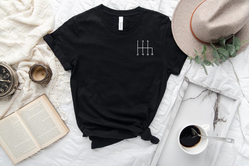 May include: Black t-shirt with a white graphic of a car's gear shift pattern. The numbers 1, 2, 3, 4, 5, and R are visible on the graphic.