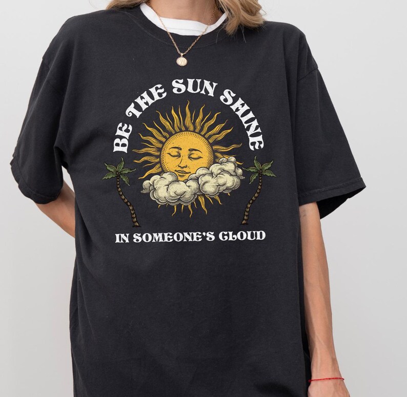 Pu&ograve; includere: Maglietta nera con una grafica bianca di un sole con un viso e il testo "Be the sun shine in someone's cloud".