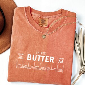 May include: A coral-coloured t-shirt with the text "SALTED BUTTER AA" printed in white. The shirt also includes a measurement guide for tablespoons and cups of butter.
